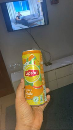 a hand holding up a can of lipton orange juice in front of a television