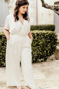 a woman in white jumpsuits and heels standing on the sidewalk with her hands behind her back