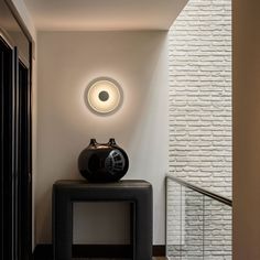 a black vase sitting on top of a table next to a white brick wall with a circular light above it