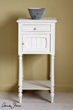 a white table with a bowl on top of it next to a wall and wooden floor