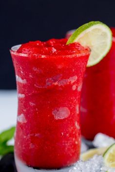 two glasses filled with red liquid next to limes and ice on a black plate