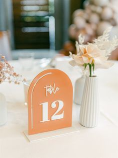 an orange table number sitting next to a white vase with flowers in it on top of a table