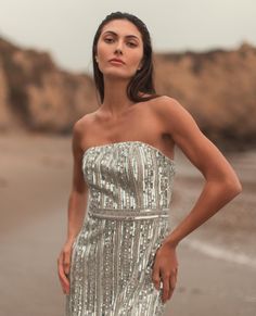 a woman is standing on the beach in a silver dress with her hands on her hips