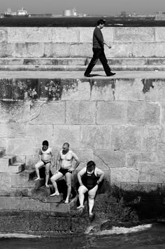 black and white photograph of four men sitting on the edge of a stone wall, with one man jumping in the air