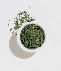 a white bowl filled with green tea leaves on top of a white tablecloth next to a pile of dried herbs