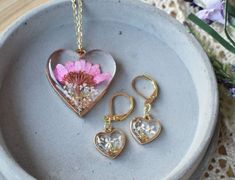 This beautiful handmade botanical jewelry set features a golden heart shaped pendant and earrings, with a purple Daisy and white Queen Anne's Lace flowers, sprouting from a reflective bed of crushed glass. Pressed Flowers that will never die,  preserved forever in crystal clear resin, with a beautiful domed finish.Flower Name & Meaning-  Daisy- new beginnings, rebirth, love. Queen Anne's Lace- safety, beauty, attract loveBezels/ Pendant Frame-  Heart shaped golden frame -1.2" wide x 1.3" tall -A Rose Gold Heart Pendant Jewelry With Flower Charm, Mother's Day Flower Shaped Jewelry With Heart Charm, Dainty Heart Pendant Jewelry With Birth Flower, Mother's Day Flower Jewelry With Heart Charm, Mother's Day Jewelry With Heart Charm And Flower Shape, Handmade Rose Gold Heart Pendant Jewelry, Mother's Day Flower Shaped Jewelry With Matching Earrings, Heart-shaped Jewelry With Flower Charm For Wedding, Nature-inspired Heart Shaped Jewelry Gift