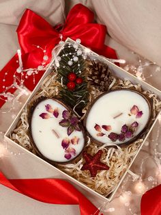 two white candles in a box with flowers and pine cones on top, surrounded by red ribbon