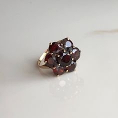 A vintage 9 carat yellow gold Garnet floral cluster ring. This beautiful piece holds a gorgeous collection of deep red stones in a gold setting. CONDITION: Good condition, wear consistent with age and use. Please see photos for more detail. HALLMARKED 9 CARAT GOLD, ASSAYED IN LONDON 1975 SETTING SIZE DIAMETER: 18mm SMALLER STONE SIZE: 6mm (.50 carat) CENTRAL STONE SIZE: 7mm (1.0 carat) BAND WIDTH: 1.5mm RING SIZE: UK: N | US: 6 3/4 WEIGHT: 3.4 grams (UUZ) Vintage Red Garnet Cluster Ring, Vintage Garnet Cluster Ring As Gift, Vintage Gold Ruby Cluster Ring, Vintage Gold Cluster Ruby Ring, Antique Cluster Ring As A Gift, Vintage Ruby Cluster Ring For Formal Occasions, Vintage Ruby Cluster Ring For Formal Events, Vintage Style Red Cluster Ring In 14k Gold, Vintage Gemstone Cluster Ring