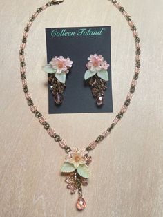 Colleen Toland Fairy Pink Flower Pendant Set.   Fairy Pink flower pendant necklace with lovely soft peach colored flowers surrounded with hand painted green leaves and a single pink fire-polish drop.  The necklace is made with delicate pink seed beads and linked together with gold colored jump-rings.  The closure is a lobster claw with a 4 inch extender so as the length can be adjusted. The earrings are made with the same lovey pink flowers and leaves and are finished with pink drop beads. Earrings and Necklaces sold separately or as a set. This is a quality piece of jewelry that will last a life time. Created by designer Colleen Toland who has been creating her unique style of vintage jewelry for more than 30 years. All of our pieces come with an engraved tag with Colleen's name so as you Handmade Pink Pendant Flower Necklace, Delicate Handmade Pink Flower Necklace, Pink Bohemian Jewelry With Handmade Flowers, Bohemian Pink Jewelry With Handmade Flowers, Elegant Handmade Peach Jewelry, Pink Beaded Flower Pendant Jewelry, Handmade Rose Gold Dangle Necklaces, Peach Flower Shaped Jewelry For Gift, Peach Flower-shaped Jewelry For Gift