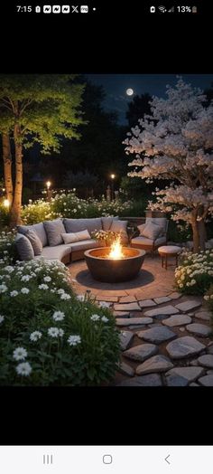 a fire pit in the middle of a stone patio surrounded by flowers and trees at night