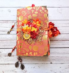 an orange and red book with flowers on it sitting on a white wooden table next to a beaded tassel