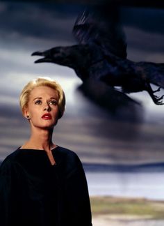 a woman standing in front of a black bird flying over her head and looking at the camera