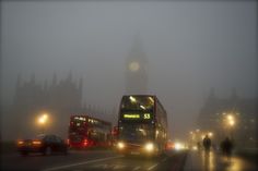 a double decker bus driving down a street in the fog with big ben in the background