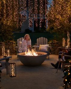 a woman sitting in front of an outdoor fire pit surrounded by christmas lights and trees