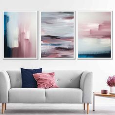 three abstract paintings hang on the wall above a couch in a white living room with pink and blue accents