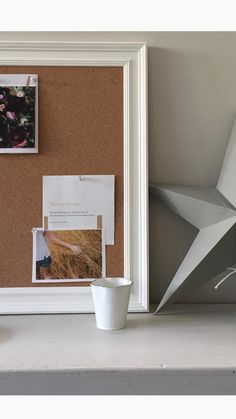 a cork board with pictures on it next to a white cup and vase filled with flowers