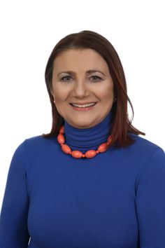 a woman wearing a blue turtle neck sweater and orange necklace smiles at the camera while standing in front of a white background