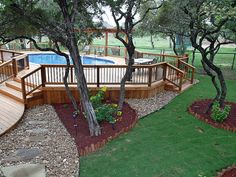 a wooden deck next to a swimming pool surrounded by trees and landscaping area with green grass