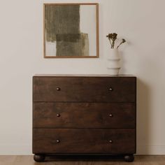 a wooden dresser sitting next to a white vase with flowers in it on top of a hard wood floor