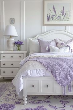 a white bed topped with purple pillows next to a night stand and dresser under a painting