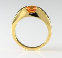 A brilliant orange gem Spessartite Garnet is bar set in a streamlined stylish 14k yellow gold ring. Spessartite, a rare form of garnet, also called mandarin or Malaya garnet, ranges from a yellow-orange through brilliant orange to orange-red. They combine very good hardness with excellent brilliance and sparkle. Spessartite is found in Africa and like all garnets is untreated. Garnet is the birthstone for January. Metal: 14k Yellow Gold The spessartite garnet is a 5.5mm round and weighs .84 cara Modern Topaz Ring With Center Stone In Yellow Gold, Modern Yellow Gold Topaz Ring With Center Stone, Modern Yellow Gold Sapphire Ring With Center Stone, Formal Orange Topaz Ring With Center Stone, Modern Gold Topaz Ring With Center Stone, Modern 14k Gold Topaz Ring For Formal Occasions, Modern Orange Rings For Gift, Modern Gold Sapphire Ring, Modern Gold Sapphire Ring With Center Stone