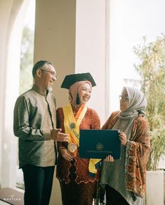 two men and a woman standing next to each other in front of a window holding a diploma