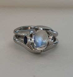 The Crystal Ball Ring is a very unique design, crafted by Heirloom Pavé in 18K white gold, featuring a Cabochon Moonstone. And two perfectly blue round brilliant Sapphires. The Moonstone is a oval shape Cabochon with a beautiful iridescent that so mystical and other worldly looking. The two, bezel-set Sapphires are a total carat weight of .35ct are a beautiful accent to the center. In the high polished white gold it is a true statement piece. Size 7 (Sizable upon request) Every Antique piece of Unique White Multi-stone Moonstone Ring, Celestial Moonstone Cabochon Jewelry, Mystical Cabochon Ring For Anniversary, Mystical Anniversary Ring With Cabochon, Fusion Style Moonstone Ring Gift, Fusion Style Oval Moonstone Ring For Anniversary, Unique Opal Ring With Moonstone, Fusion Style Oval Moonstone Ring Gift, Fusion Style Moonstone Gemstone Ring For Anniversary