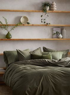 a bed with green sheets and pillows in front of wooden shelves filled with potted plants