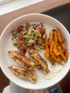 a white plate topped with chicken, fries and salad next to a window sill