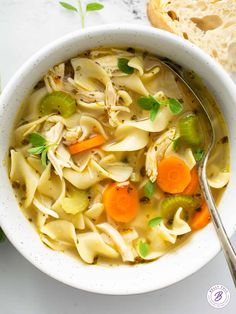 a white bowl filled with chicken noodle soup next to a slice of bread on a table