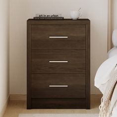 a night stand with three drawers and a cup on it next to a white bed