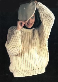 a woman in a white sweater and hat smiles while holding her hands up to her head