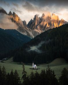 the mountains are covered in mist and clouds as the sun sets over them, with a small church at the bottom