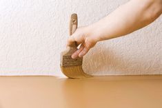 a hand holding a paint brush on top of a wooden table next to a white wall