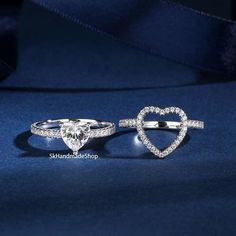 two heart shaped rings with diamonds on them sitting on a blue cloth, one in the shape of a heart