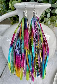 two pairs of multicolored feather earrings on a white vase with greenery in the background