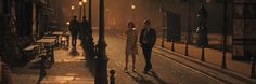 two people walking down the street at night in an old fashioned city with lights on