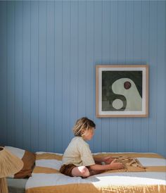 a little boy sitting on top of a bed next to a wall with a painting