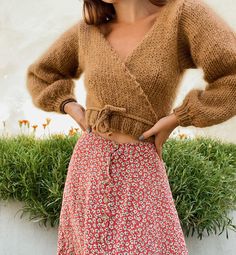 a woman standing in front of a plant wearing a brown sweater and floral print skirt