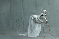 a woman with crutches and a white dress is leaning against a concrete wall