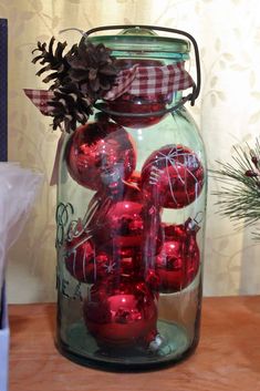 a glass jar filled with red ornaments on top of a wooden table next to a pine cone