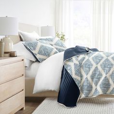 a bed with white and blue sheets in a bedroom next to a dresser, lamp and window