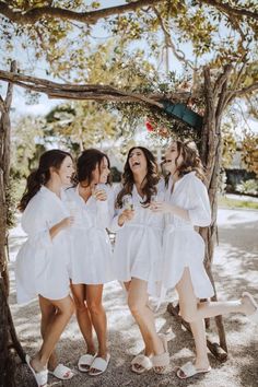 four women in white robes laughing and drinking wine under a wooden arch with branches on either side