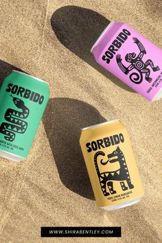 three different types of sodas sitting in the sand with their shadows on the ground