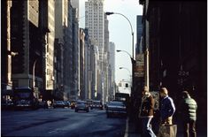 people are standing on the sidewalk in front of tall buildings and cars driving down the street