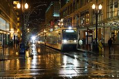 a city street at night with people walking on the sidewalk and a train coming down the track