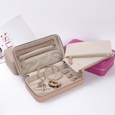 an open suitcase with jewelry inside on a white surface next to a pink box and book