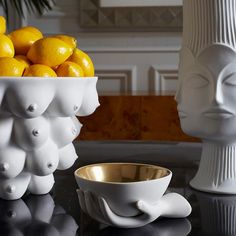 a white bowl filled with lemons next to a statue head and bowl of fruit