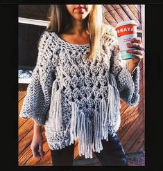 a woman is holding a book and wearing a crocheted sweater with tassels