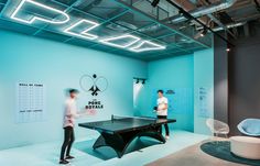 two people playing ping pong in an office setting with neon lights on the ceiling